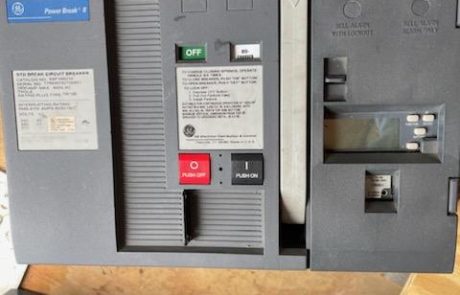 A GE Power Break II circuit breaker with a black and gray casing, placed on a wooden surface. The breaker has a red "Push Off" button and a black "Push On" button, along with a green "Off" indicator. Various labels and warnings are visible, including a specifications label on the left side. The breaker appears to be in a used or refurbished condition. In the background, other electrical components are partially visible.