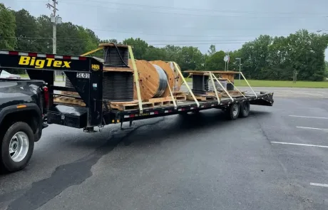 Pickup truck towing a flatbed trailer loaded with industrial pipes and a large wooden spool.